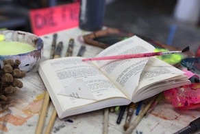 Offenliegendes Buch auf einem mit Farbflecken übersäten Arbeitstisch im Atelier. Ein rosafarbener Pinsel liegt quer über den Seiten, umgeben von Pinseln, Farbbechern und leuchtenden Neonpigmenten. Die Szene vermittelt eine Verbindung von theoretischer Auseinandersetzung und praktischer, künstlerischer Arbeit.